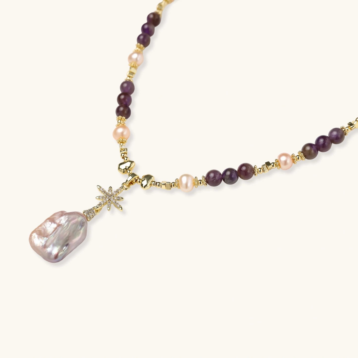 Gold necklace with iridescent baroque pearl pendant, purple amethyst beads, and crystal star charm on cream background